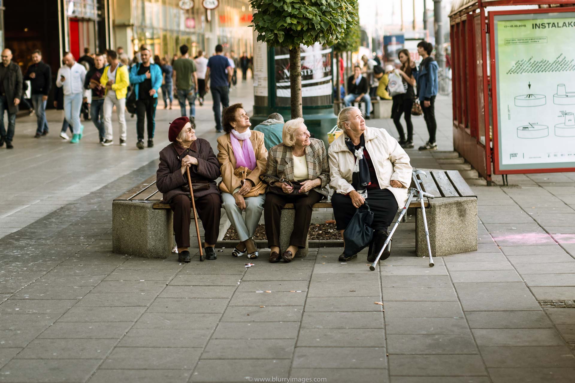 bench, four women, old woman, outdoors, 2015, bus station, pavement, blond hair, brown hat, pink scarf, white jacket, brown jacket, grey jacket, brown jacket,