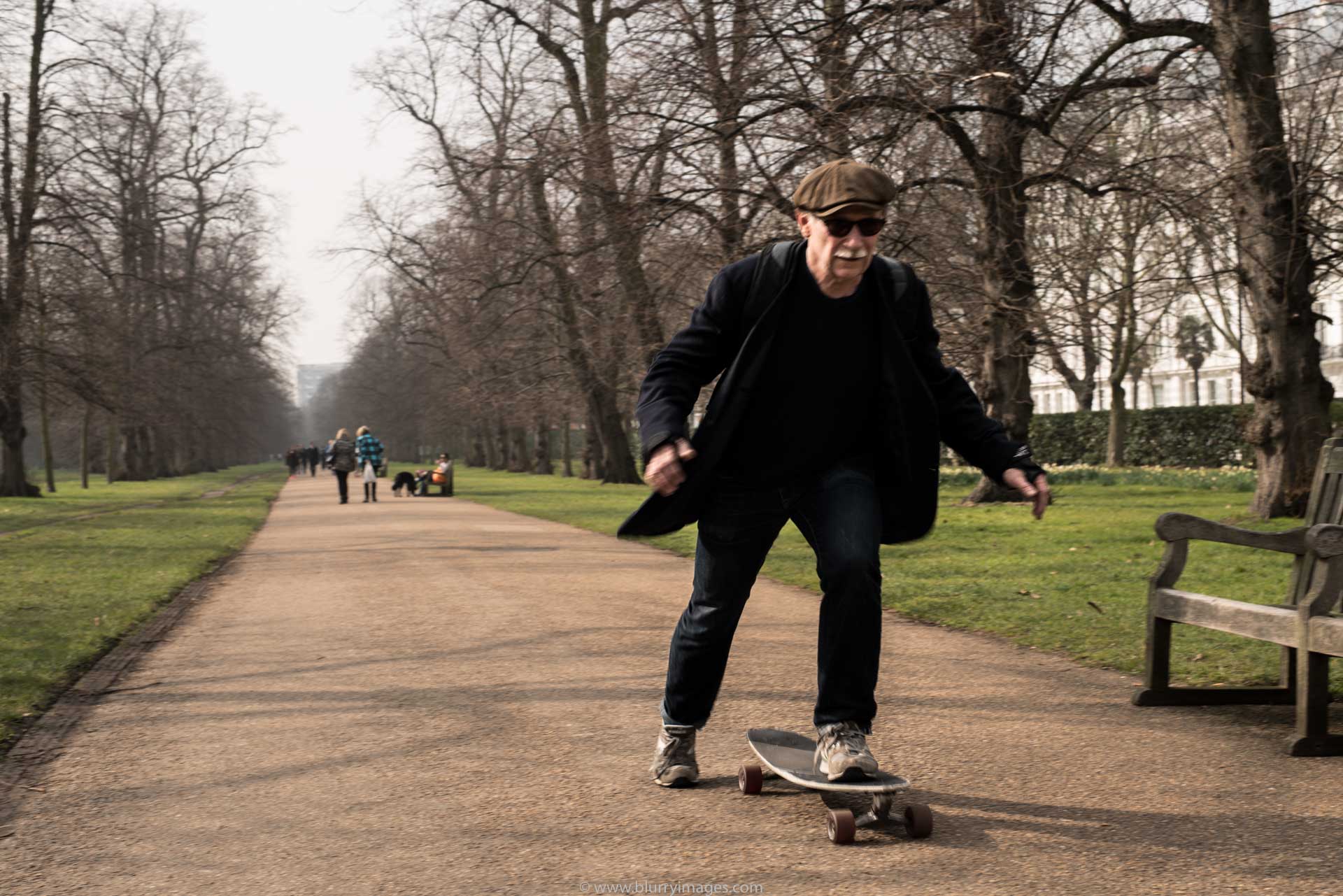 Man on a skateboard, grey hat, black glasses, grass, no leaves, black trousers, back jacket, black sweater, bench, pavement, pavement in a park , riding on a skateboard