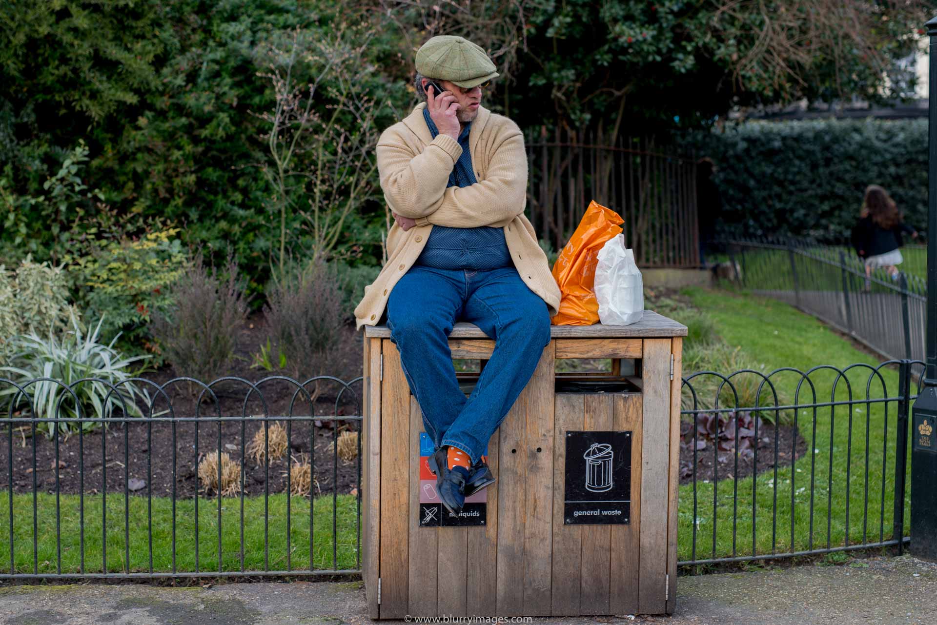 Man sitting, rubbish box, orange bag, white bag, green hat, grey blazer, orange socks, black shoes, wooden box, black fence, grass, blues jeans, outdoors, 2016, tree, green background,