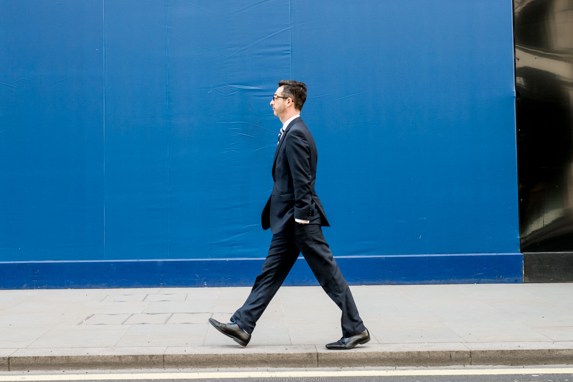 walking man, Regent Street, black suit, black shoes, pavement, man on pavement, black hair, black glasses, blue background, black background, yellow line,